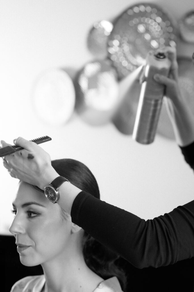 Make-Up Artist sprüht Haarspray aus einer silbernen Sprühflasche auf die dunklen Haare der Braut. Diese trägt eine glatte gestylte Frisur und einen Seidenmantel.