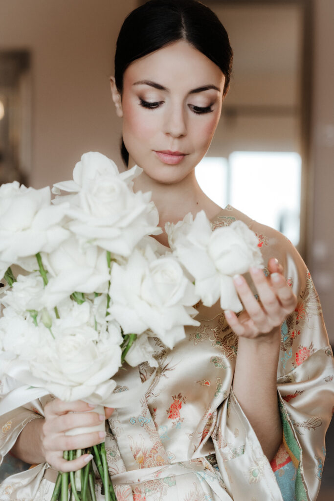 Eine wunderschöne Braut mit dunklen Augen, dunklen Haaren und eine glatt nach hinten gebundenen Frisur. Sie hält einen weißen Rosenstrauß in der Hand den sie zart berührt und auf den sie heraubschaut.